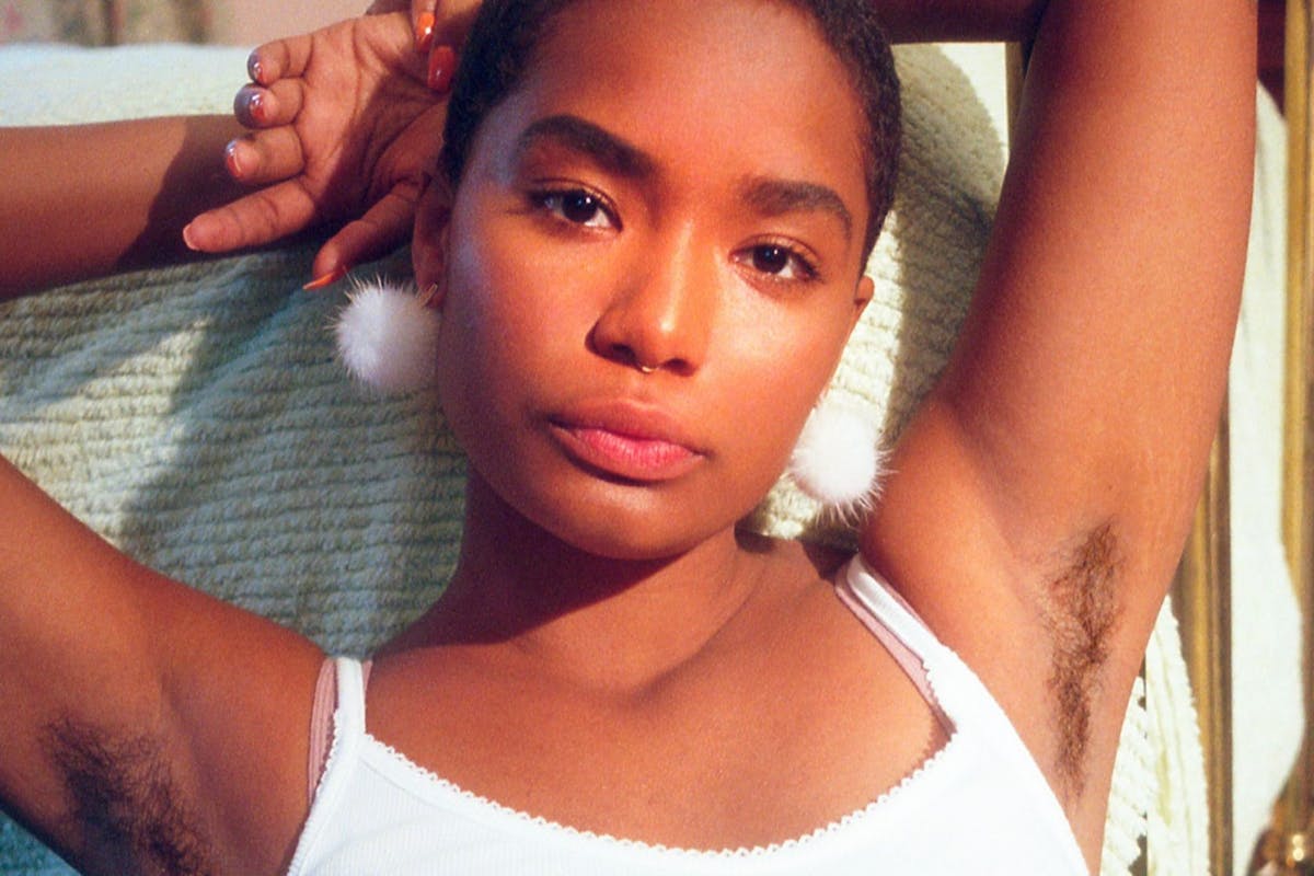 A black model dressed in a white top holds her arms behind her head with hair clearly visible under her armpits