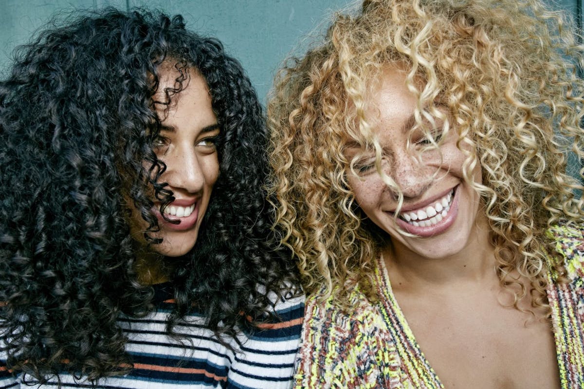 Two women with curly hair laughing. The woman on the left has black hair and the woman on the right has blonde hair.