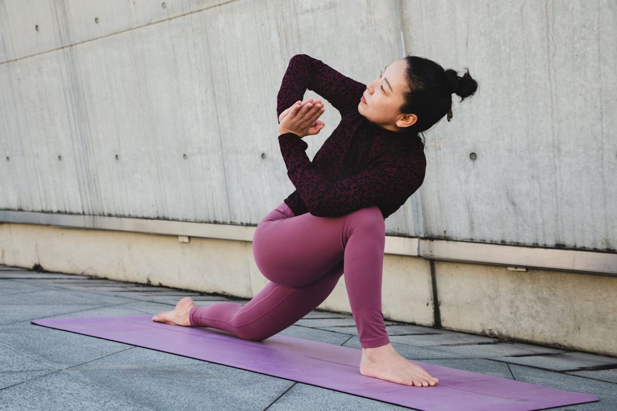 A woman doing low lunge and prayer pose in yoga on a mat outside.