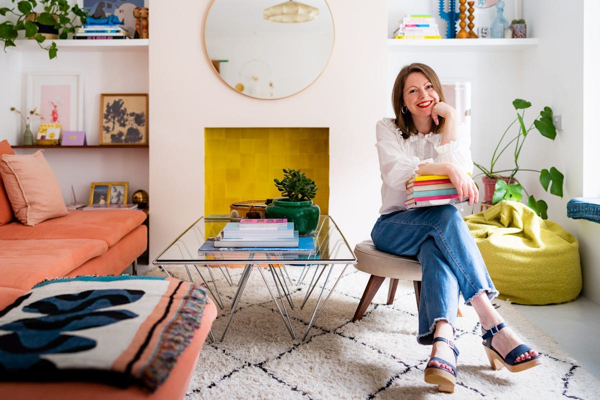 Liz Beardsel in pastel living room wearing white blouse and blue jeans and holding pile of books