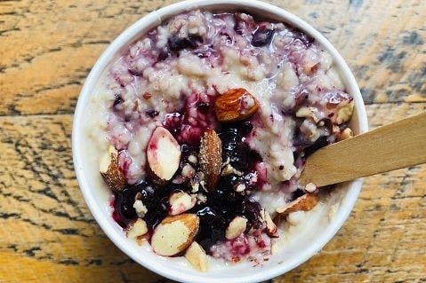 Close up of a disposable takeaway pot with vegan porridge made with coconut milk and 5 grains (oatmeal, jumbo oats, barley, rye and quinoa flakes), and berry compote and crushed almonds on a wooden table