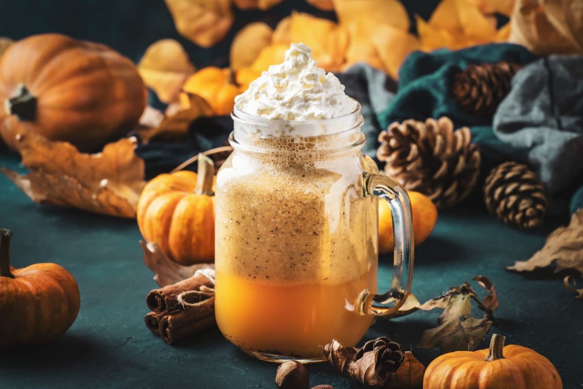 Pumpkin spiced latte or coffee in glass jar on blue table. Autumn or winter hot drink in festive natural table setting with orange leaves, spices, small pumkins, pine cones