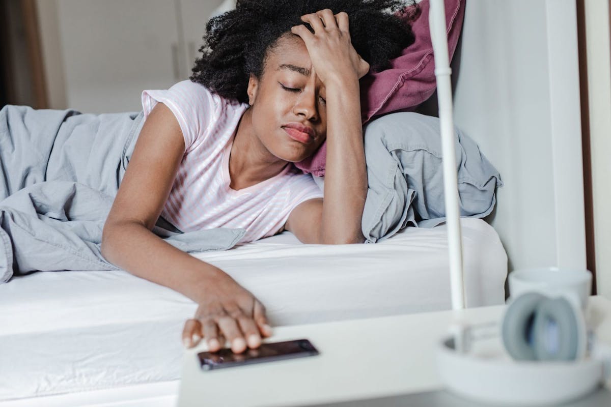 A woman with a headache reaching for her phone to turn off her morning alarm