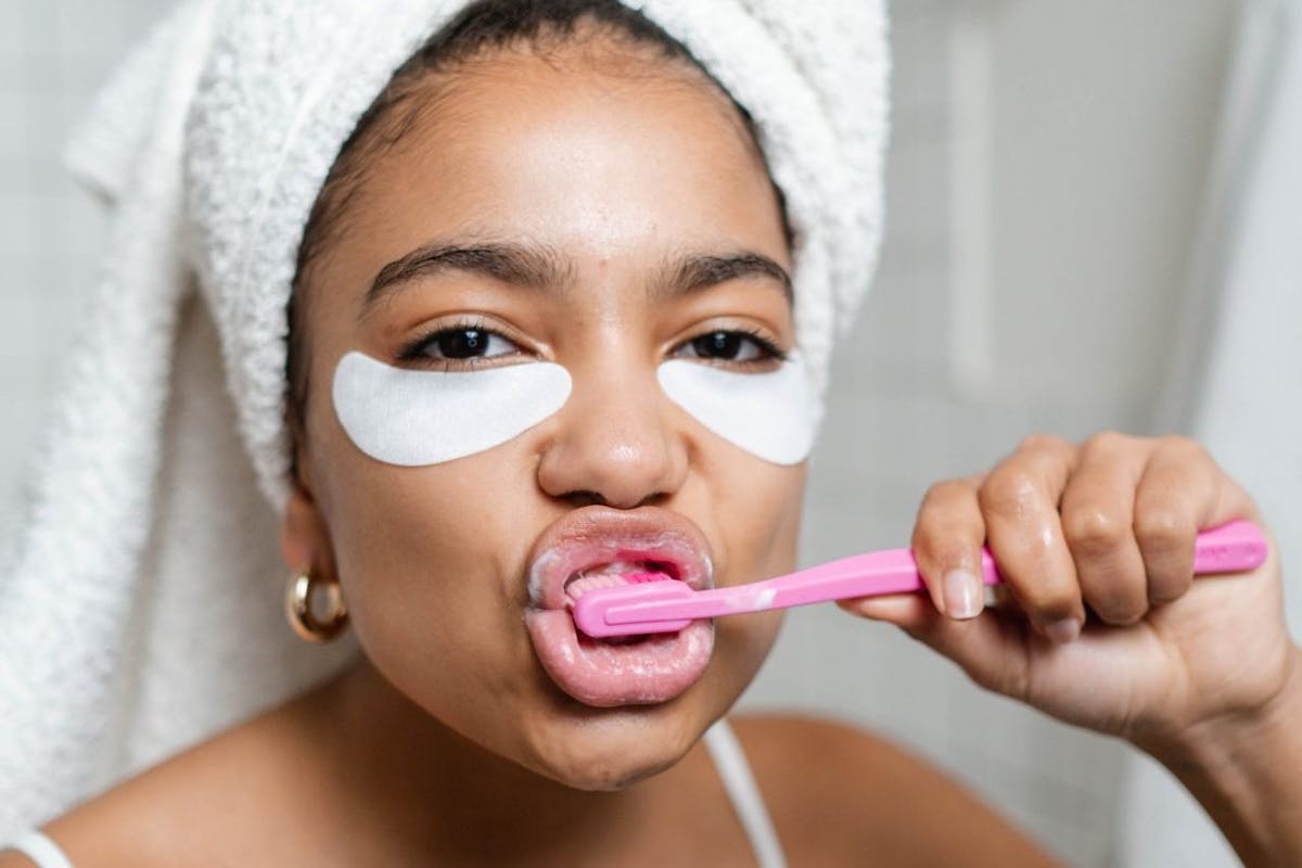 A woman with eye masks on brushing her teeth in the mirror