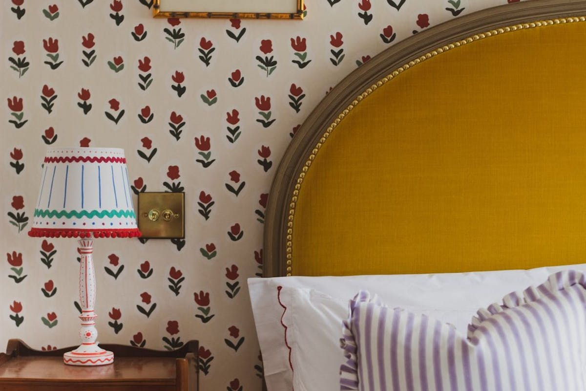 An image of the Tulip Room at Glebe house. The walls a covered with cream wallpaper that features a red, blue and green floral pattern. There is a bed with a large mustard velvet headboard and a dark wood side table with a colourful lamp